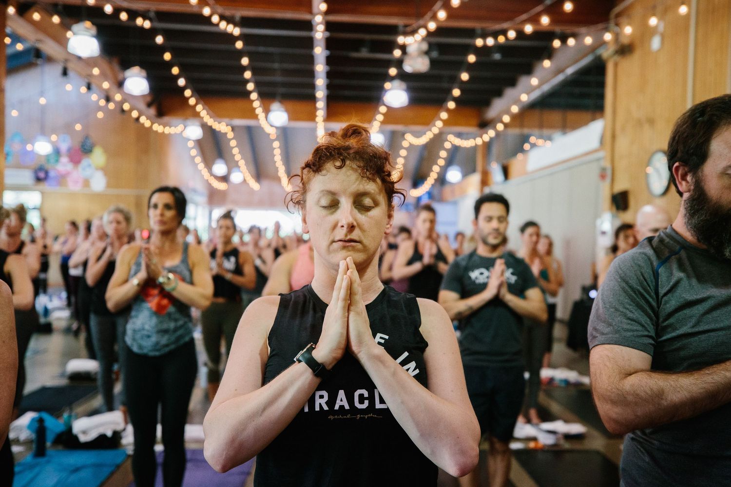 Group of people practicing yoga in a studio with string lights overhead, showing focused meditation poses.