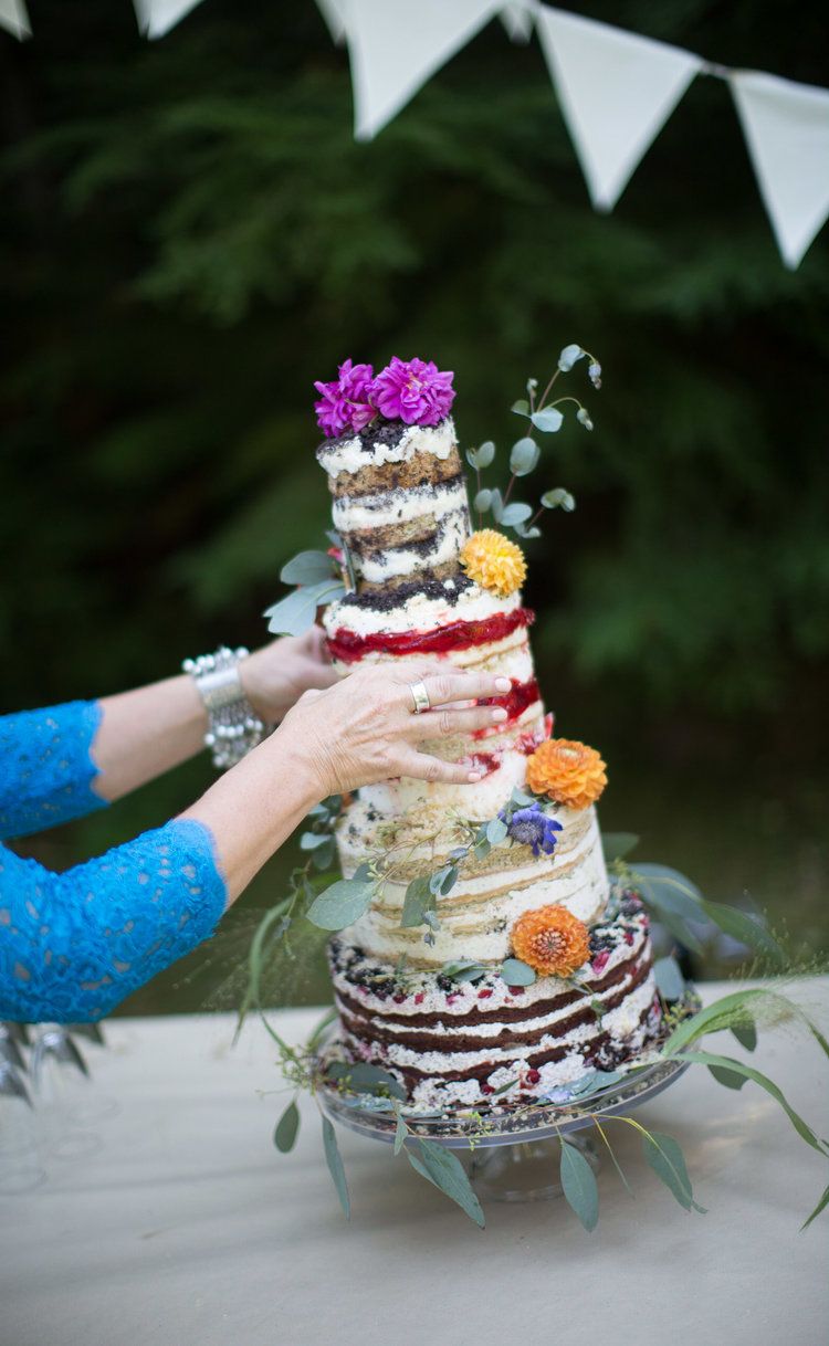 Rustic naked wedding cake decorated with fresh flowers and eucalyptus being assembled by hands in blue sleeves.