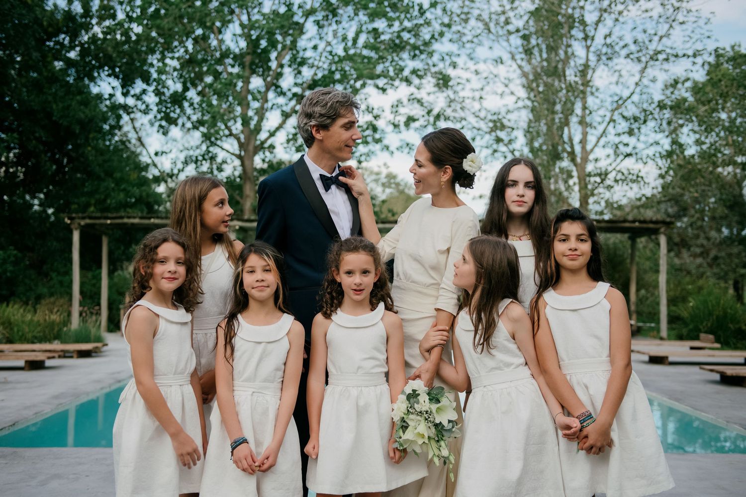 Wedding party in white dresses and formal attire stands together outdoors in front of swimming pool and greenery.