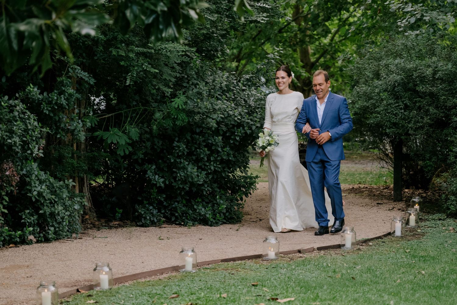 Bride and groom walk along a scenic garden path surrounded by lush green trees on their wedding day.