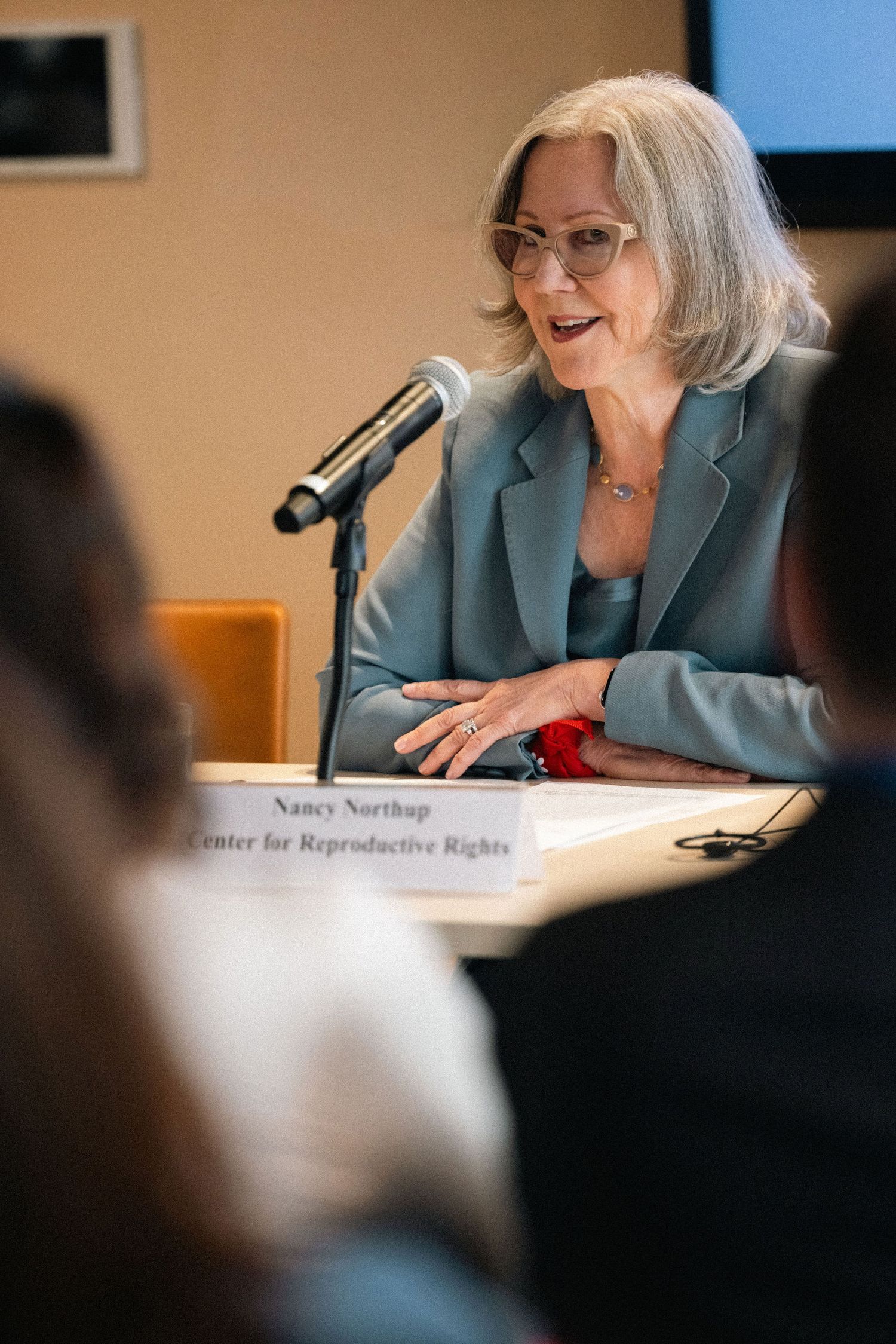 A speaker in a teal blazer and glasses smiles while speaking at a podium during a panel discussion.