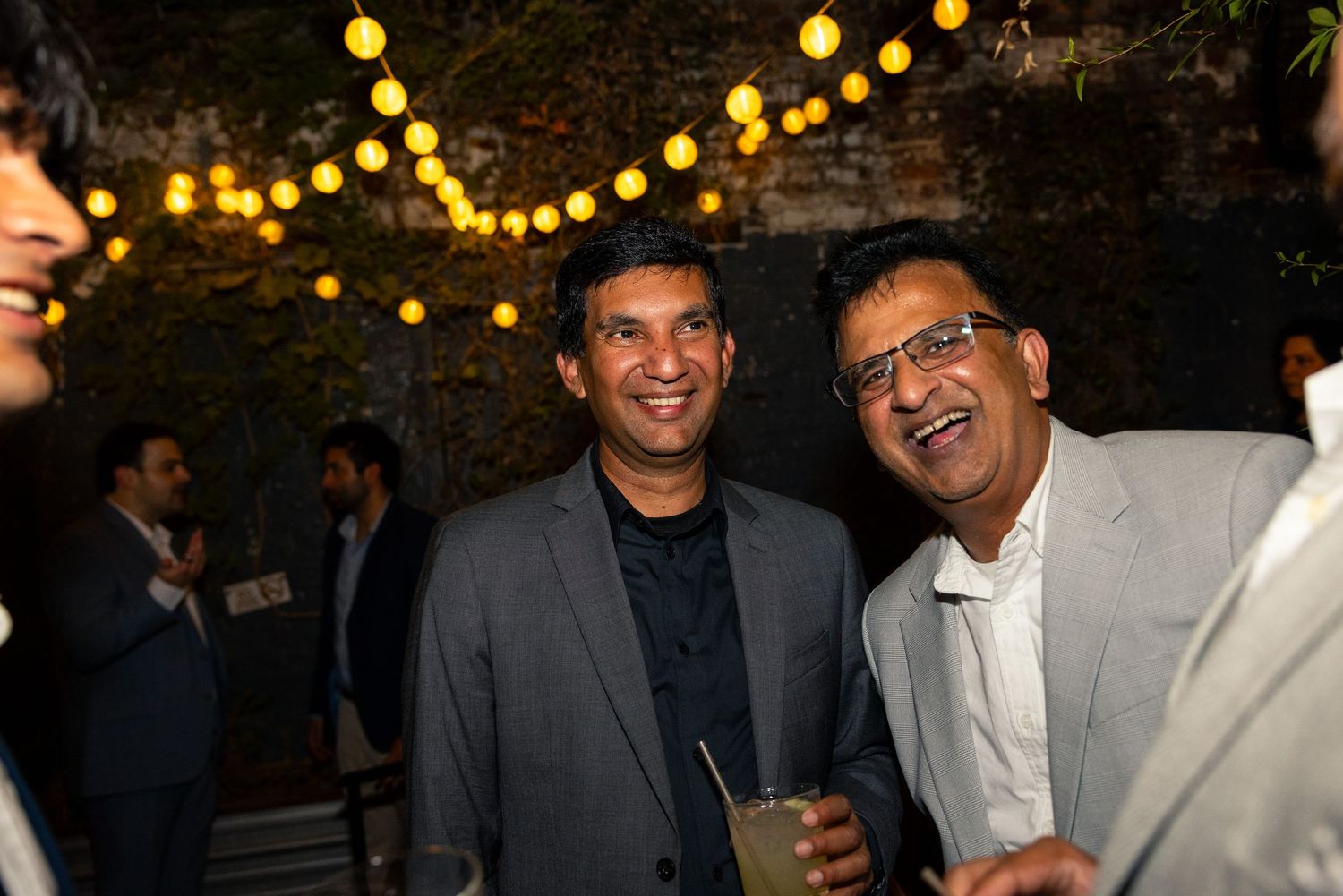 Party guests laugh and enjoy themselves under warm string lights at an outdoor evening celebration.
