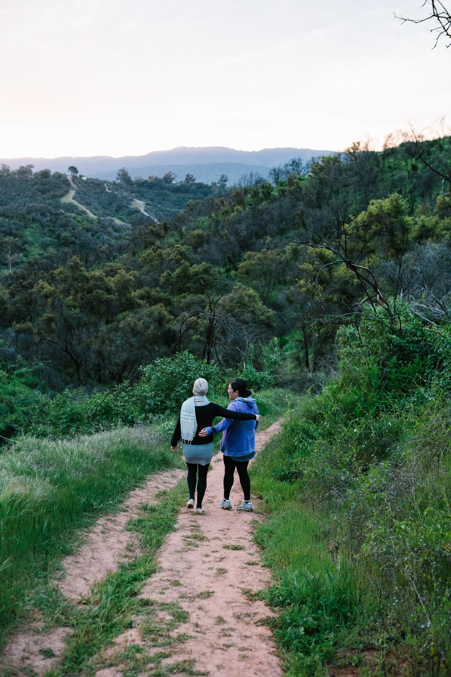 People walking on a dirt hiking trail surrounded by lush green hills and vegetation.