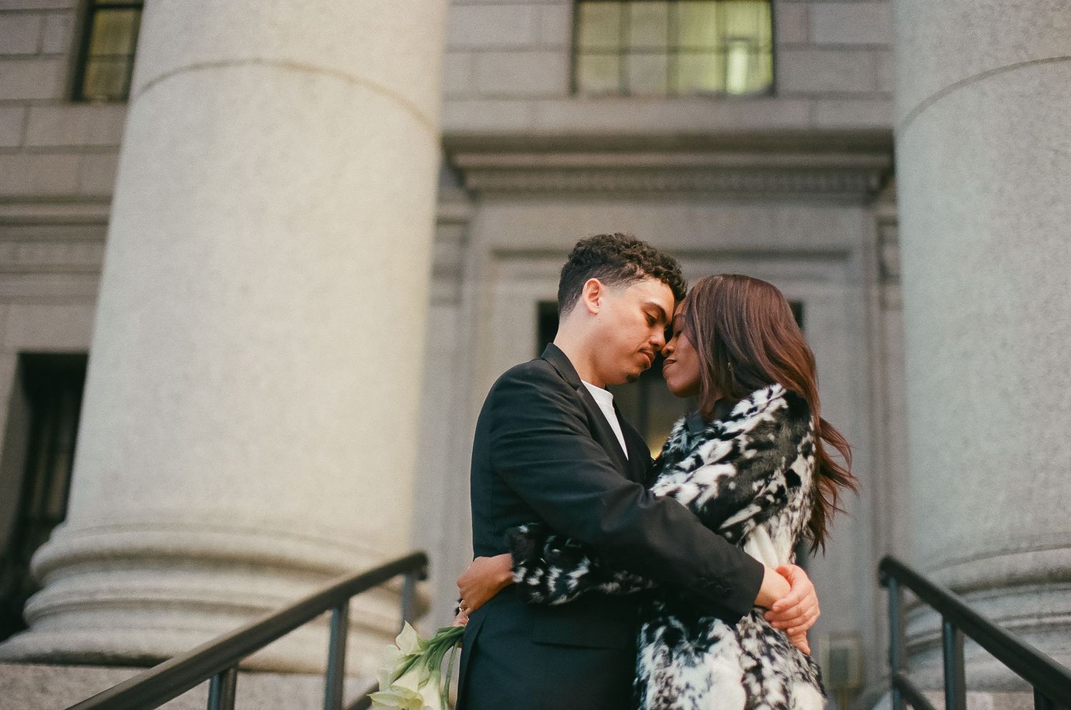 Couple sharing a romantic embrace on stone steps in front of classical columns at an outdoor engagement photo session.