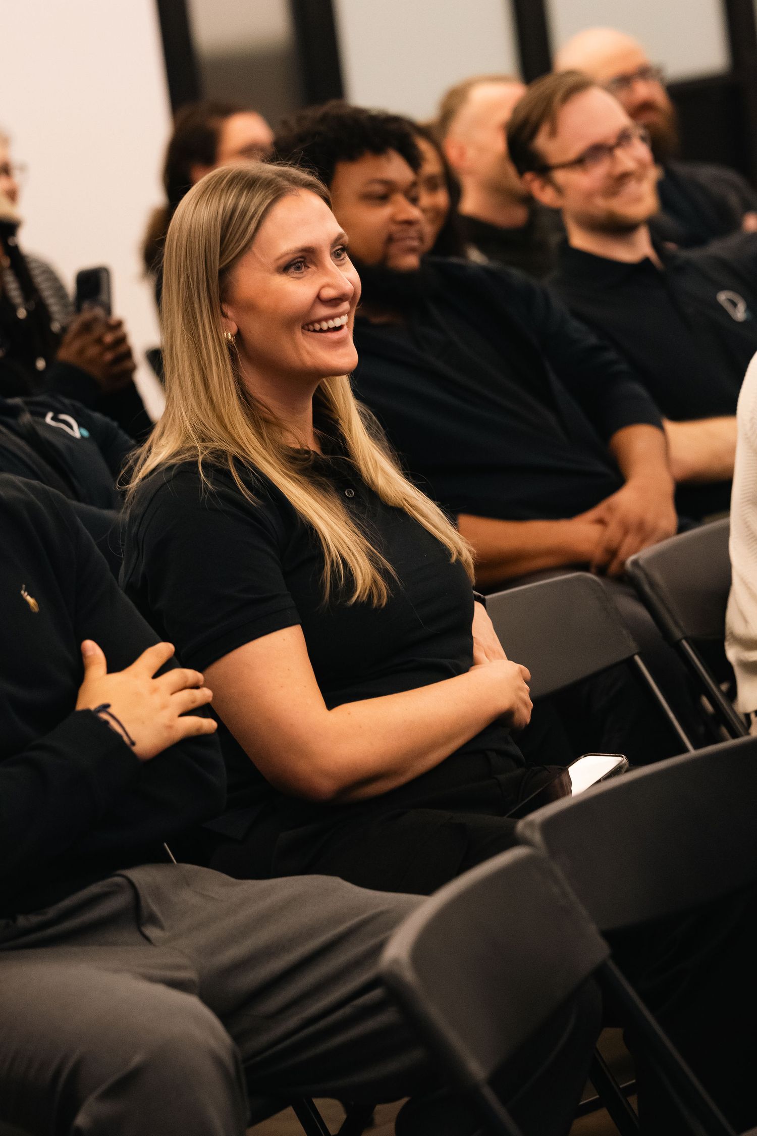 Audience members in black attire laugh and smile while seated during an engaging presentation or performance.