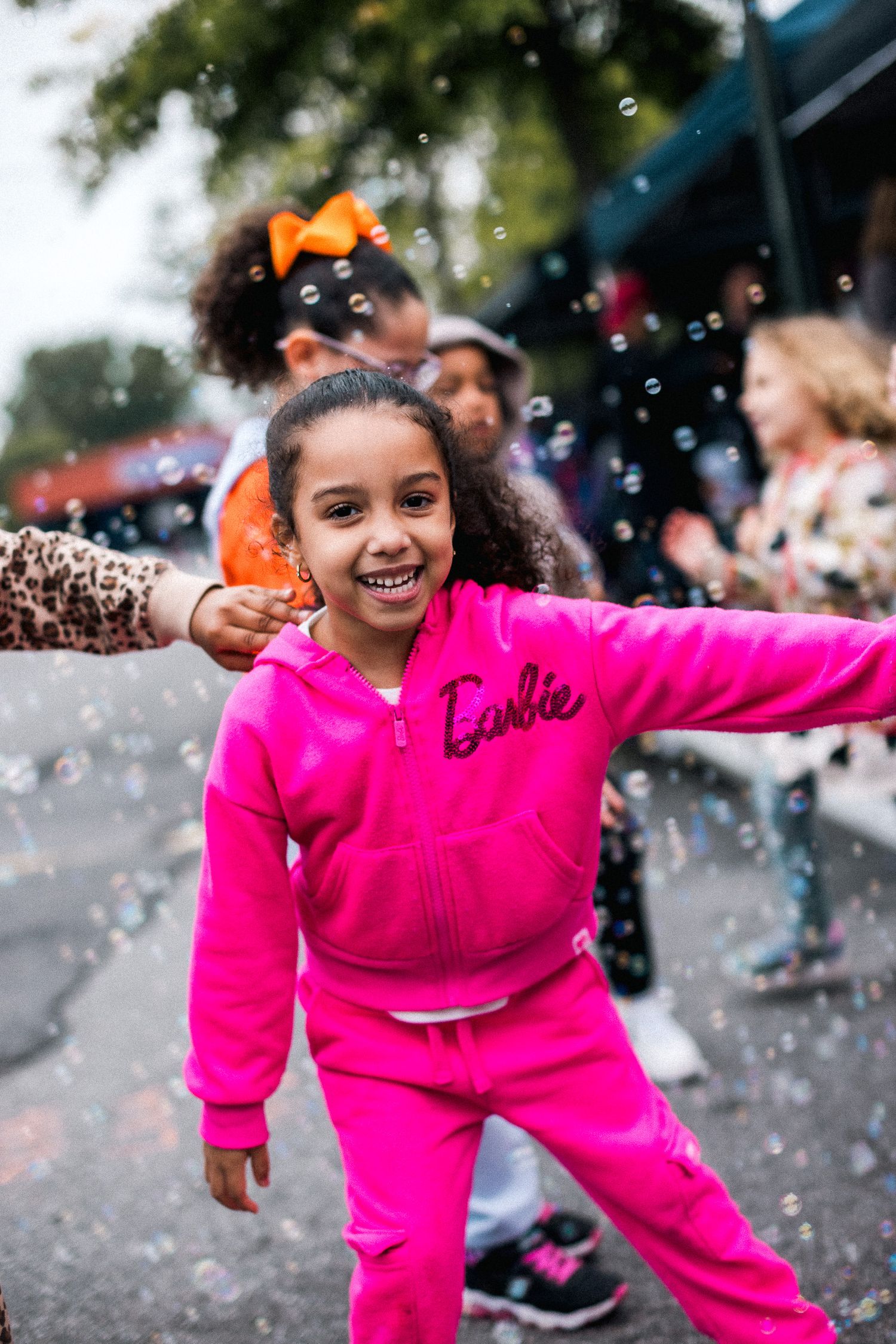 Child in bright pink Barbie outfit playing joyfully among bubbles at an outdoor celebration.