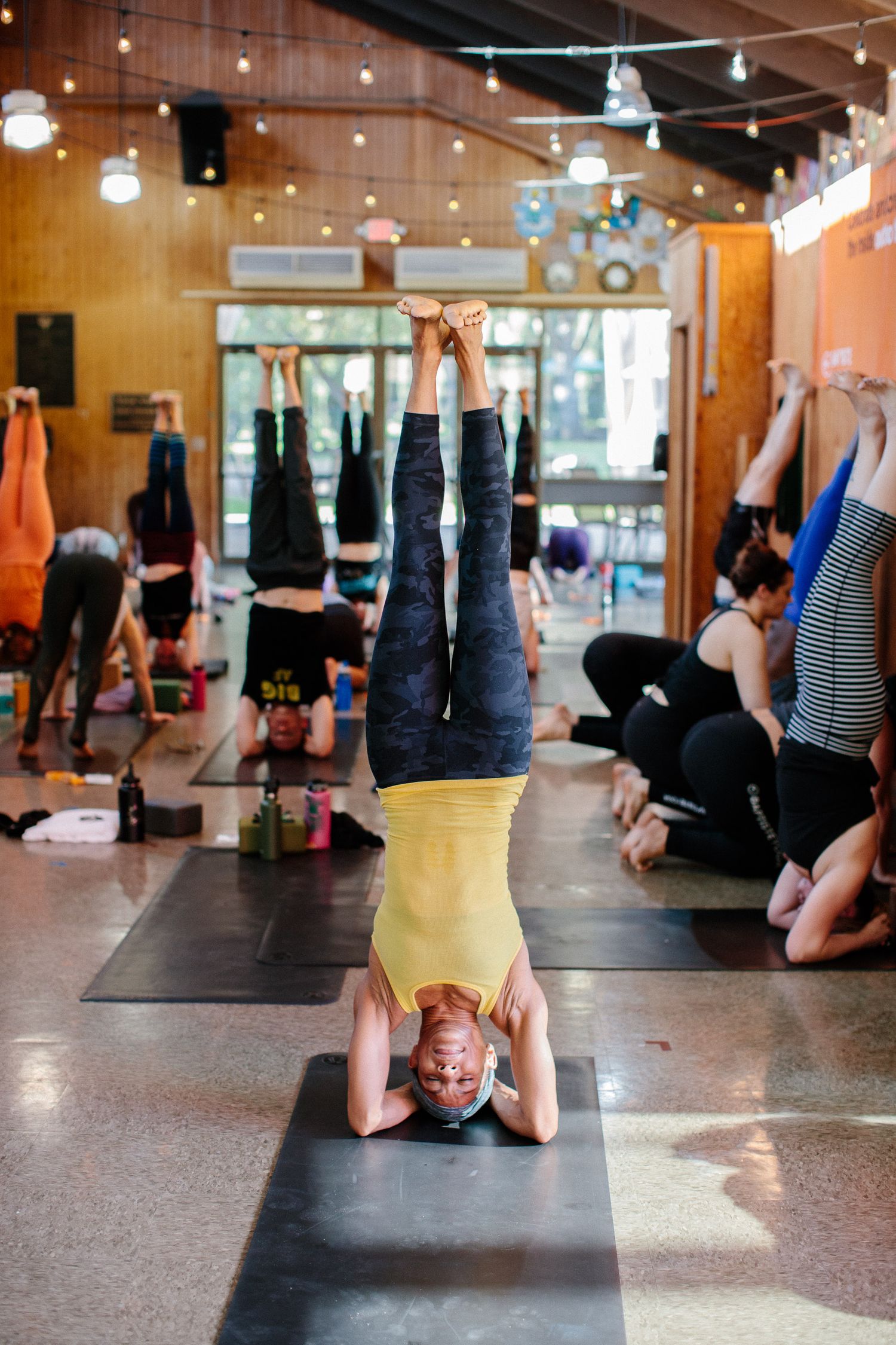 Students practicing shoulder stands in a yoga studio with string lights and wooden walls.