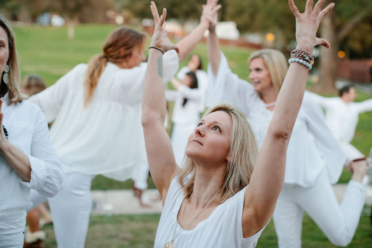 A group of participants wearing white clothing raise their arms in unison during an outdoor wellness session.