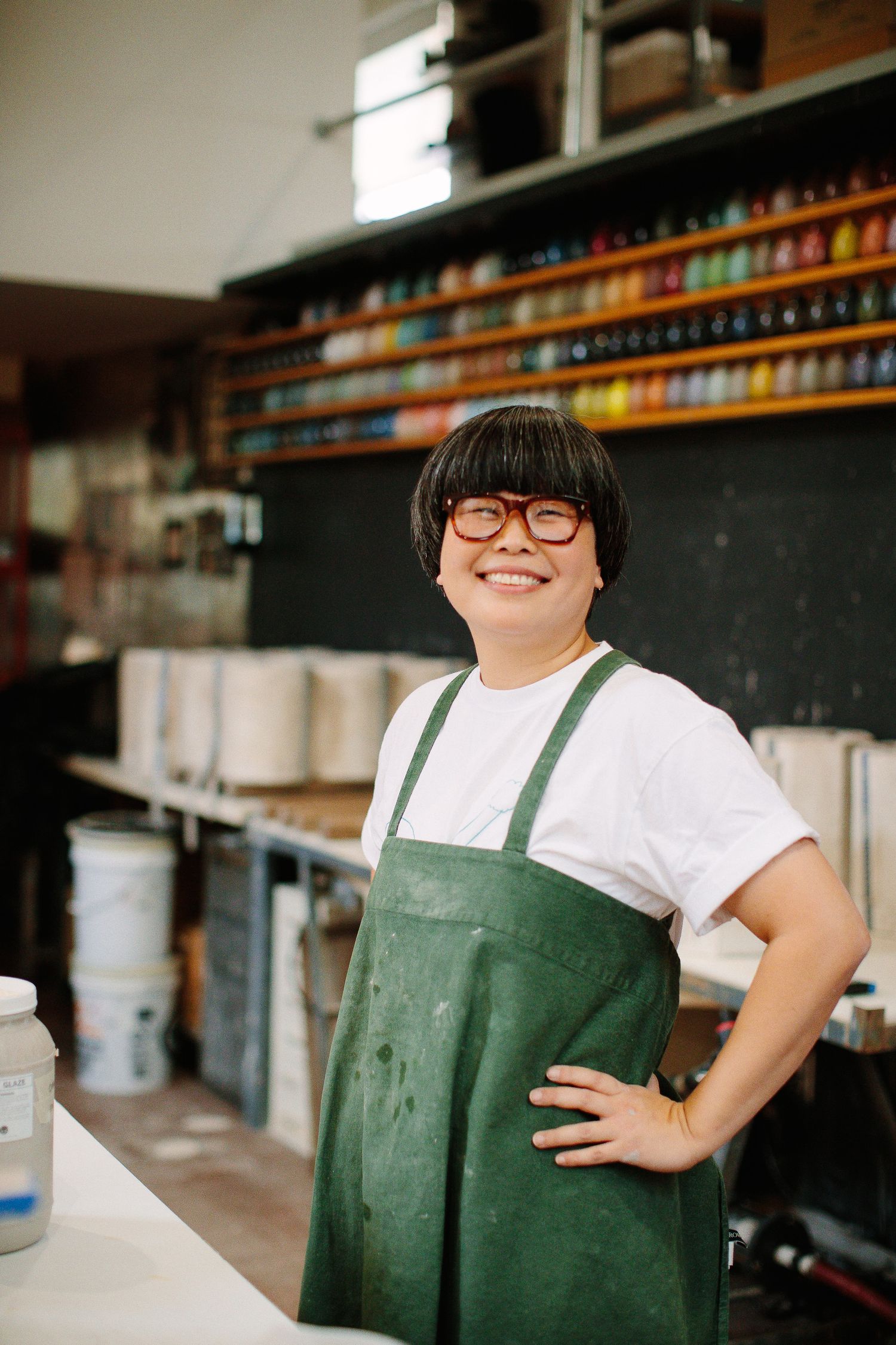Smiling person in white t-shirt and green apron stands against wall of colorful bottles in retail environment.