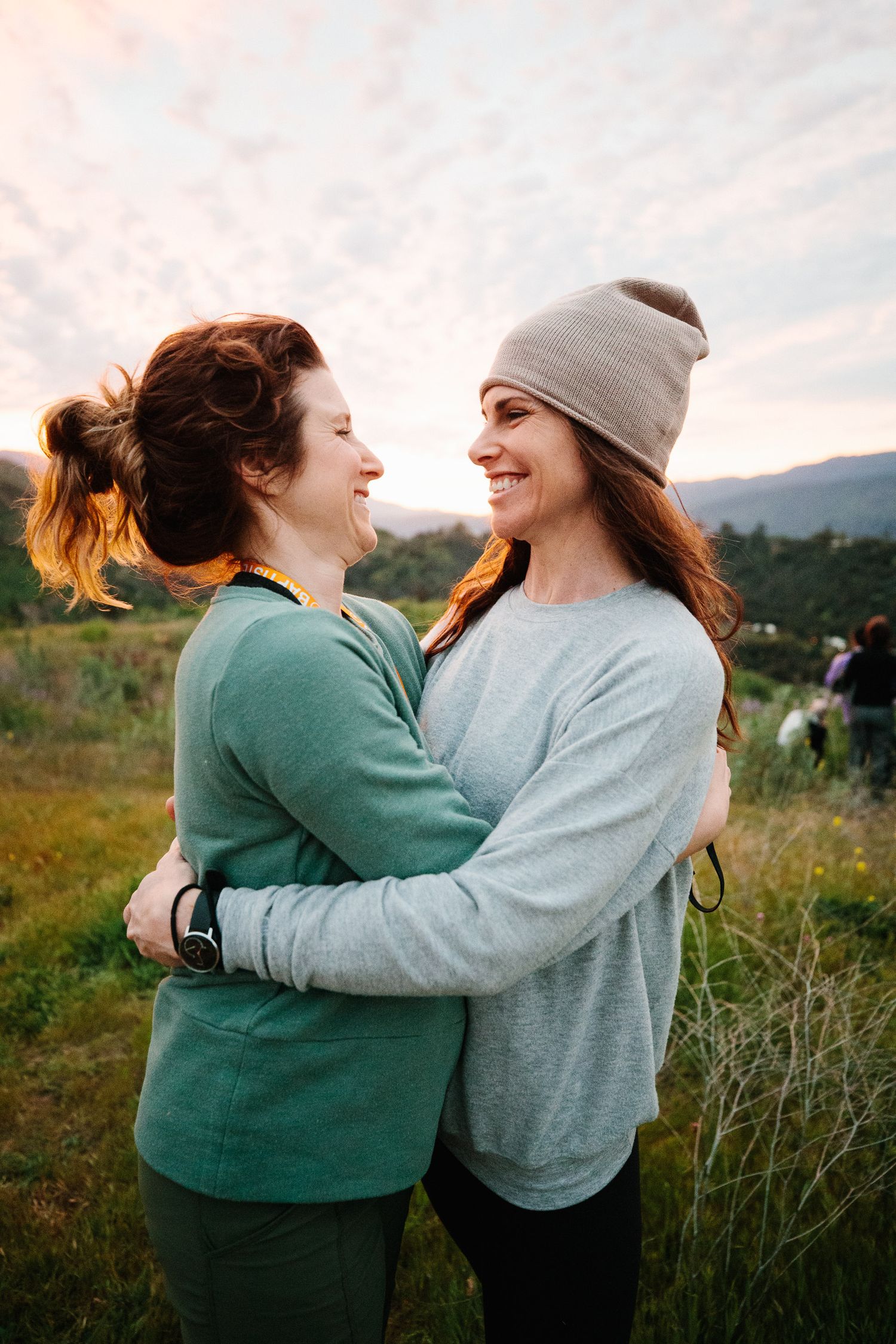 A couple embraces while laughing together in a scenic outdoor field with mountains in the background.