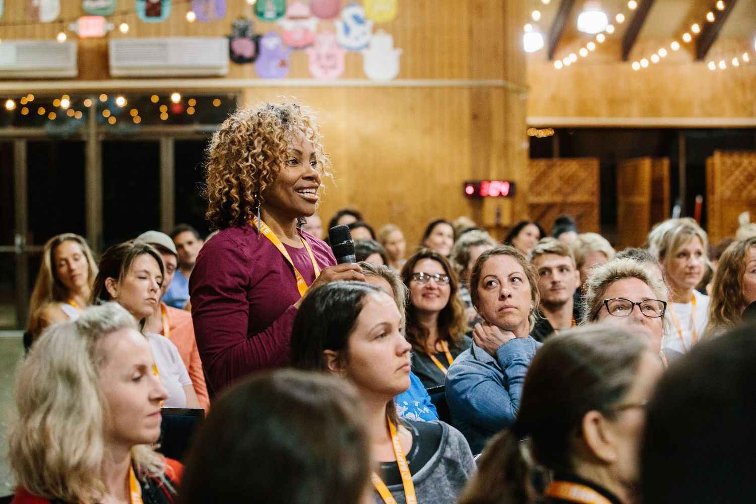 Speaker addresses attentive audience members during indoor event in wood-paneled venue with festive decorations.