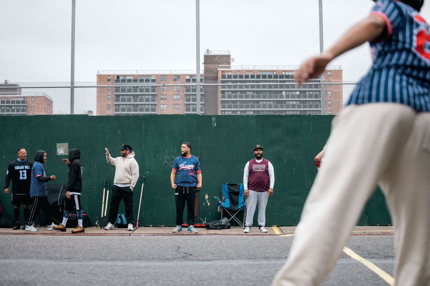 A group of people standing along a green wall while someone plays stickball in an urban setting.