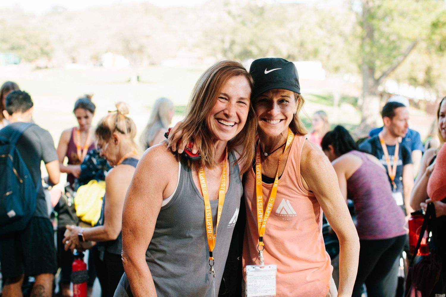 Two athletic runners wearing race medals and tank tops smile together at an outdoor sporting event in sunny weather.