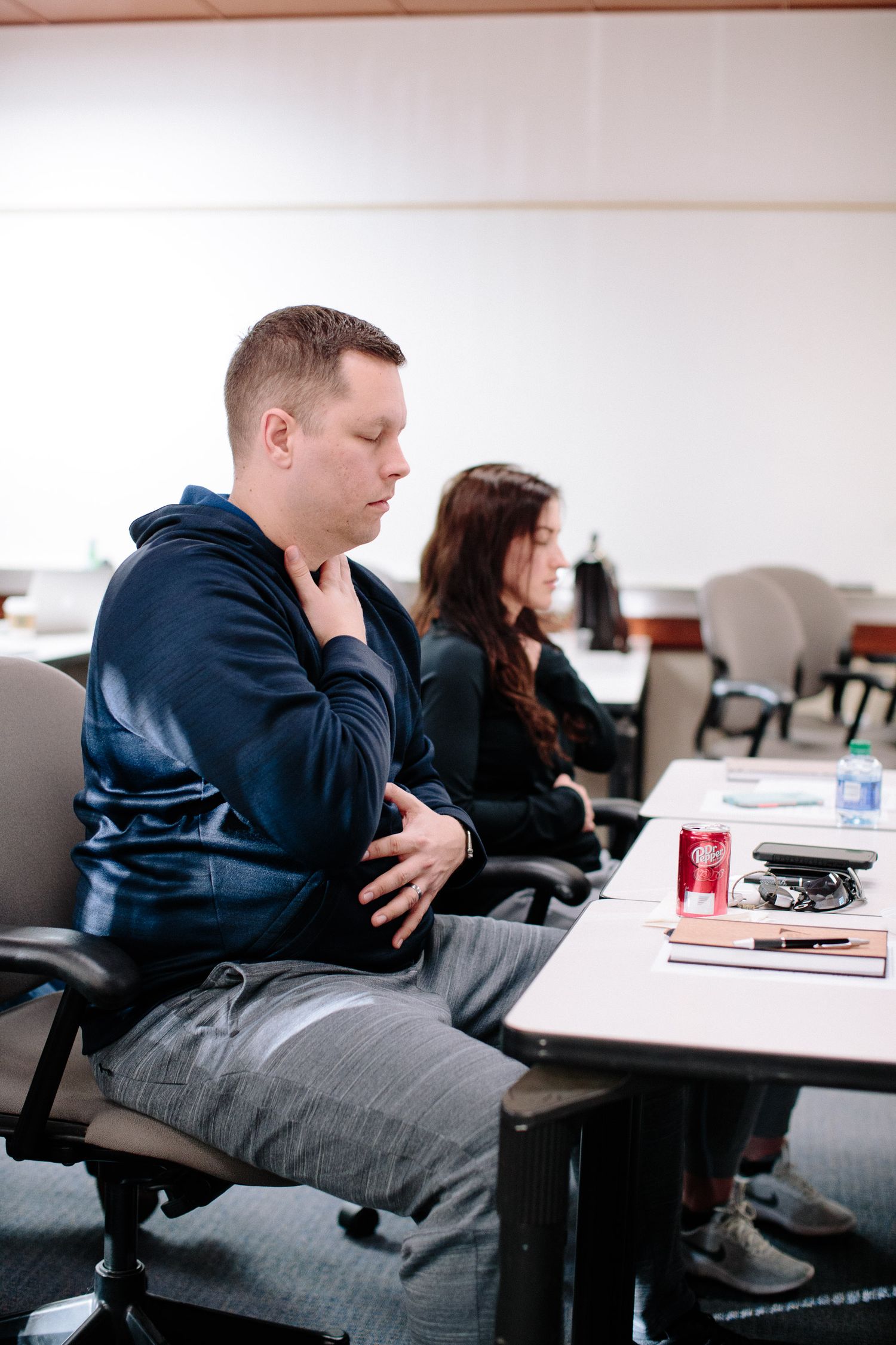 Students take notes during a classroom session with a man in a navy hoodie seated at a desk.