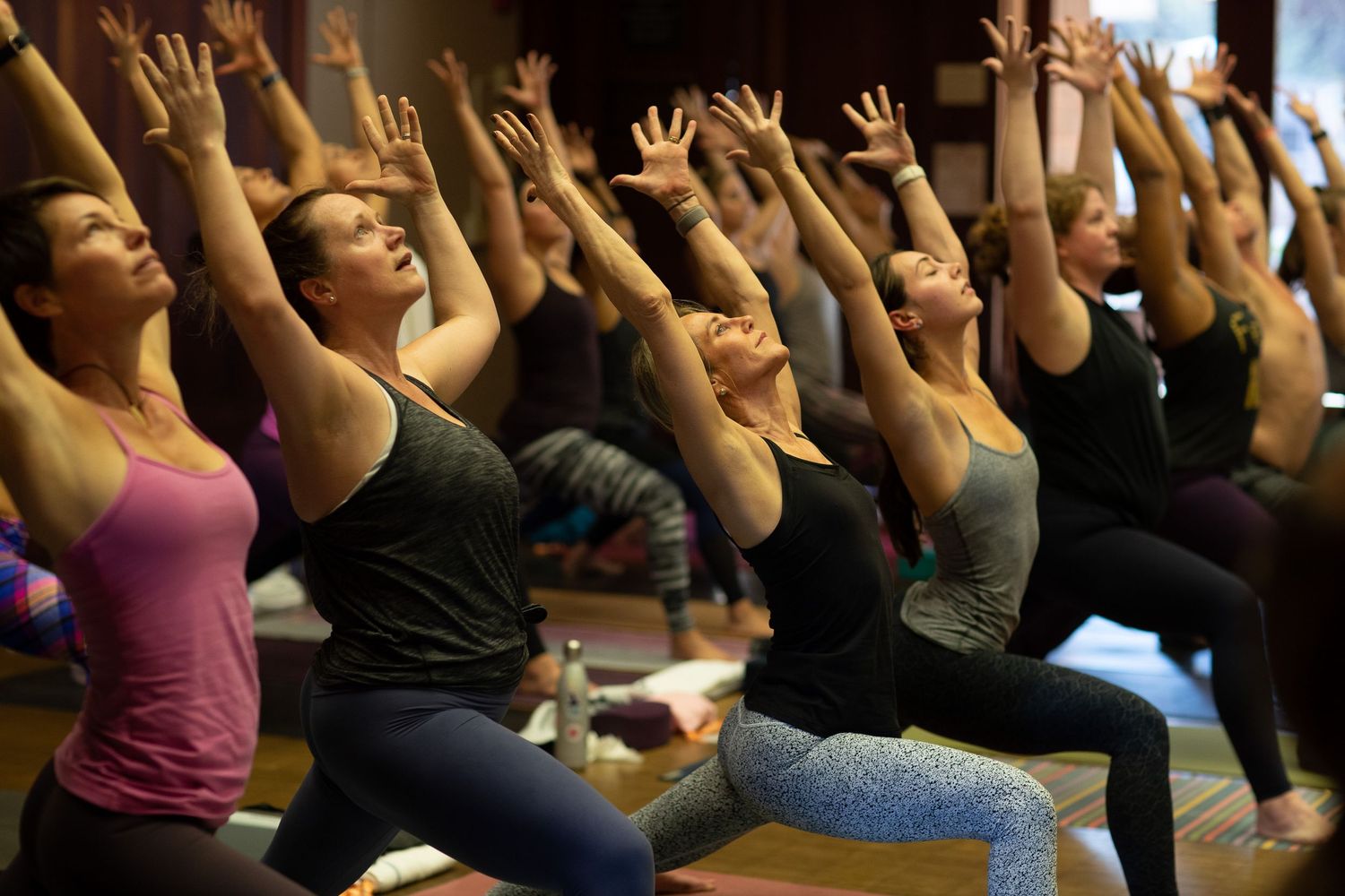 Group of yoga practitioners in warrior pose with arms raised during class.