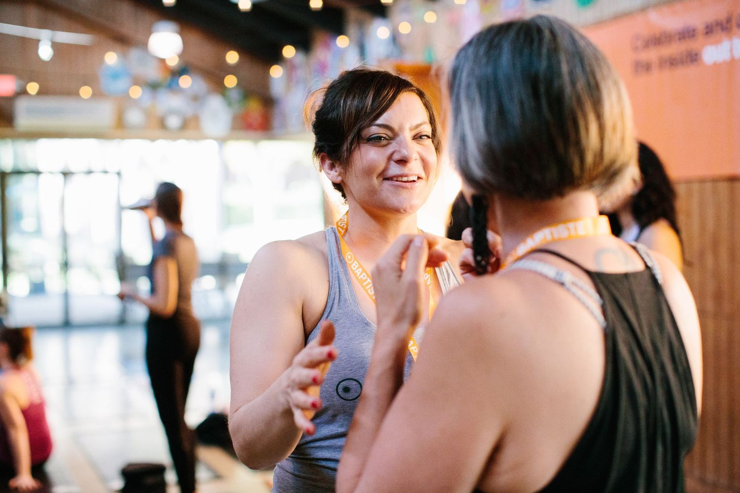Two friends share an animated conversation in a warmly lit indoor space with string lights.