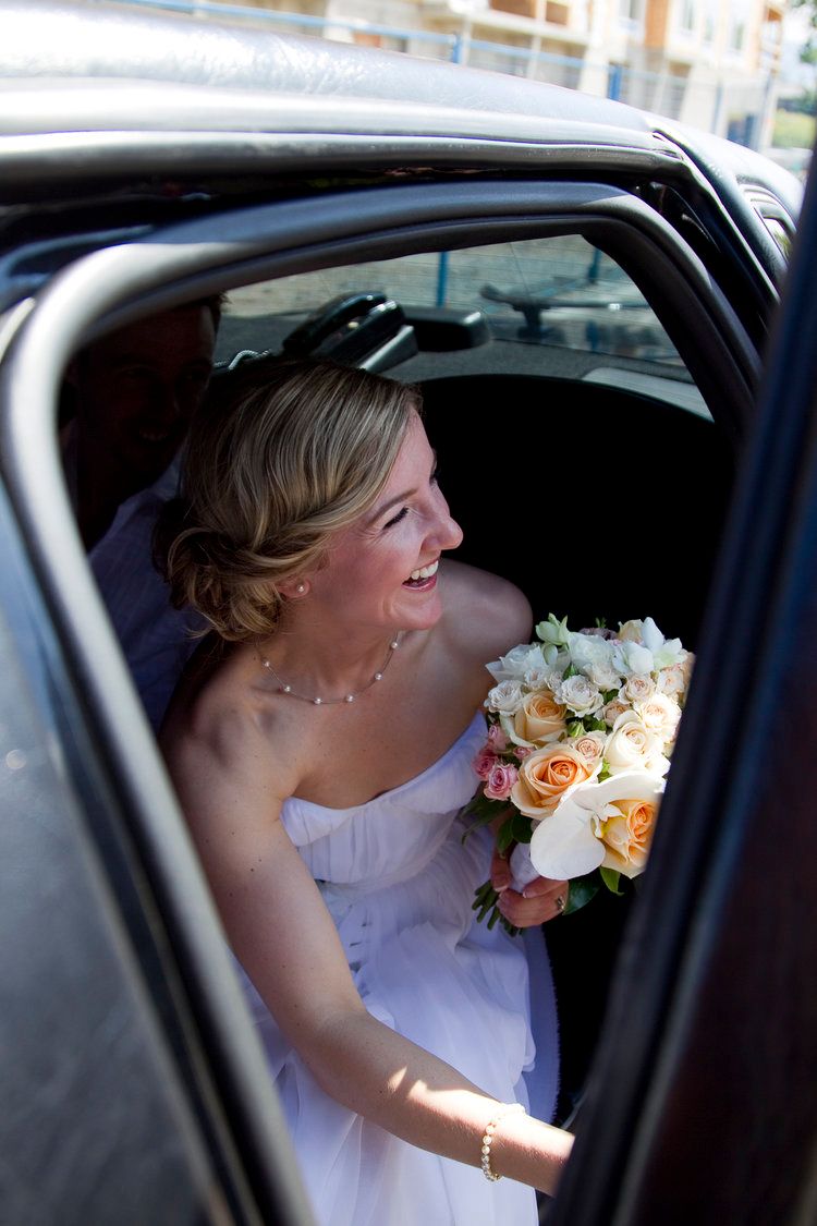 A bride in a white dress sits in a car holding a bouquet of peach and white flowers during a wedding day.