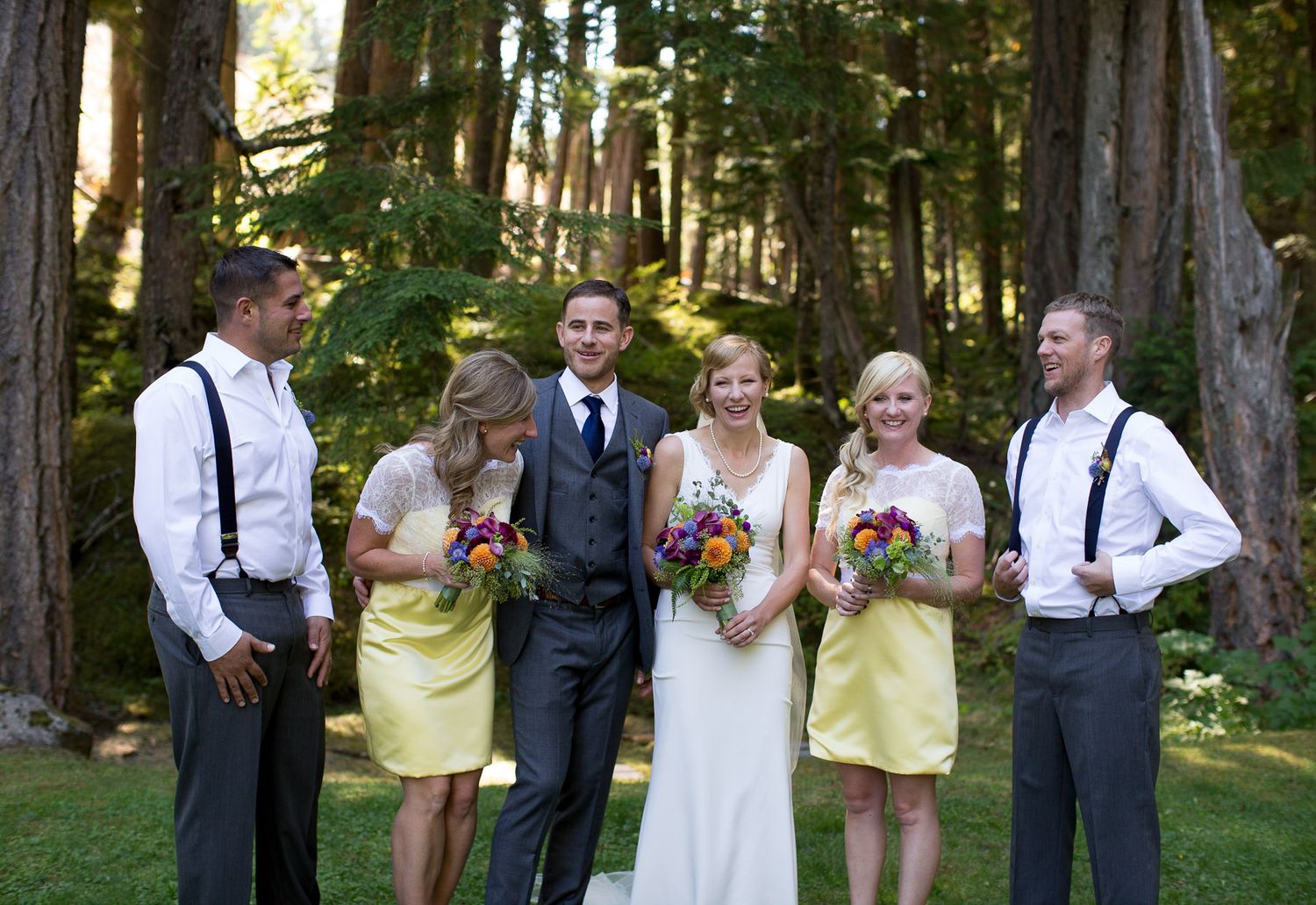 Wedding party poses in yellow dresses and suspenders against a forest backdrop.