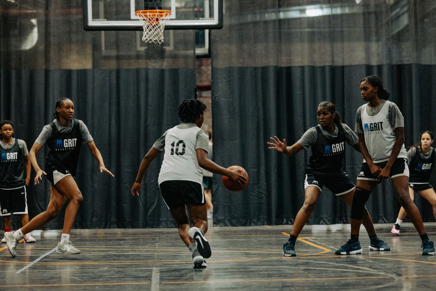 Basketball players compete in an indoor game on a dark court with a basketball hoop visible in the background.