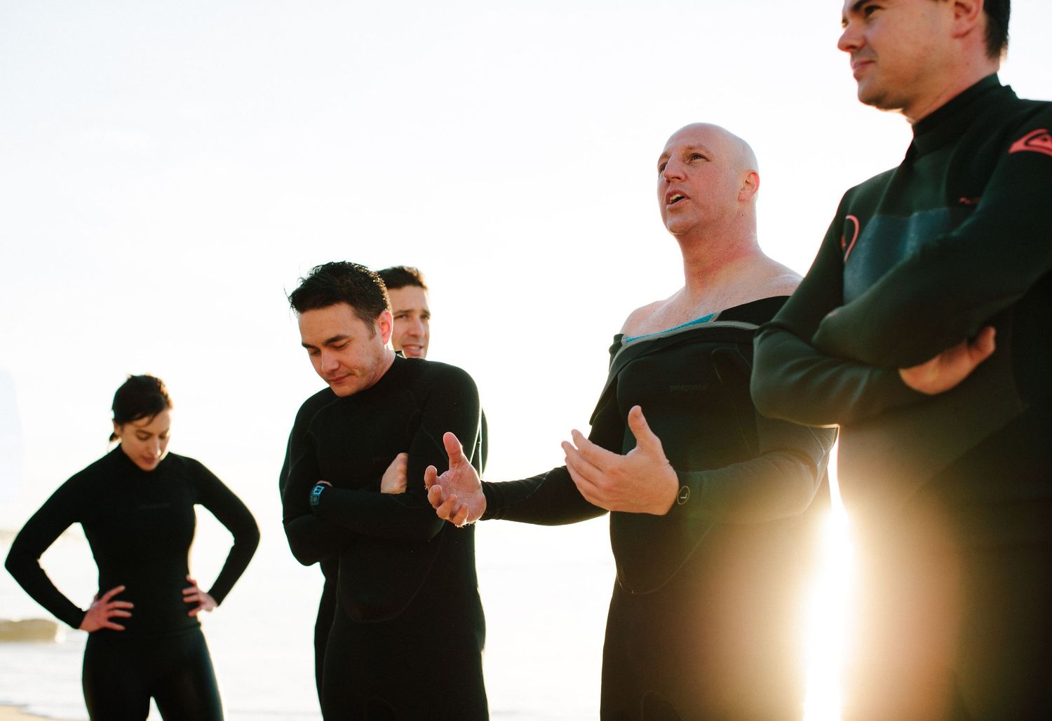 Group of people in black wetsuits standing together backlit by sunlight.