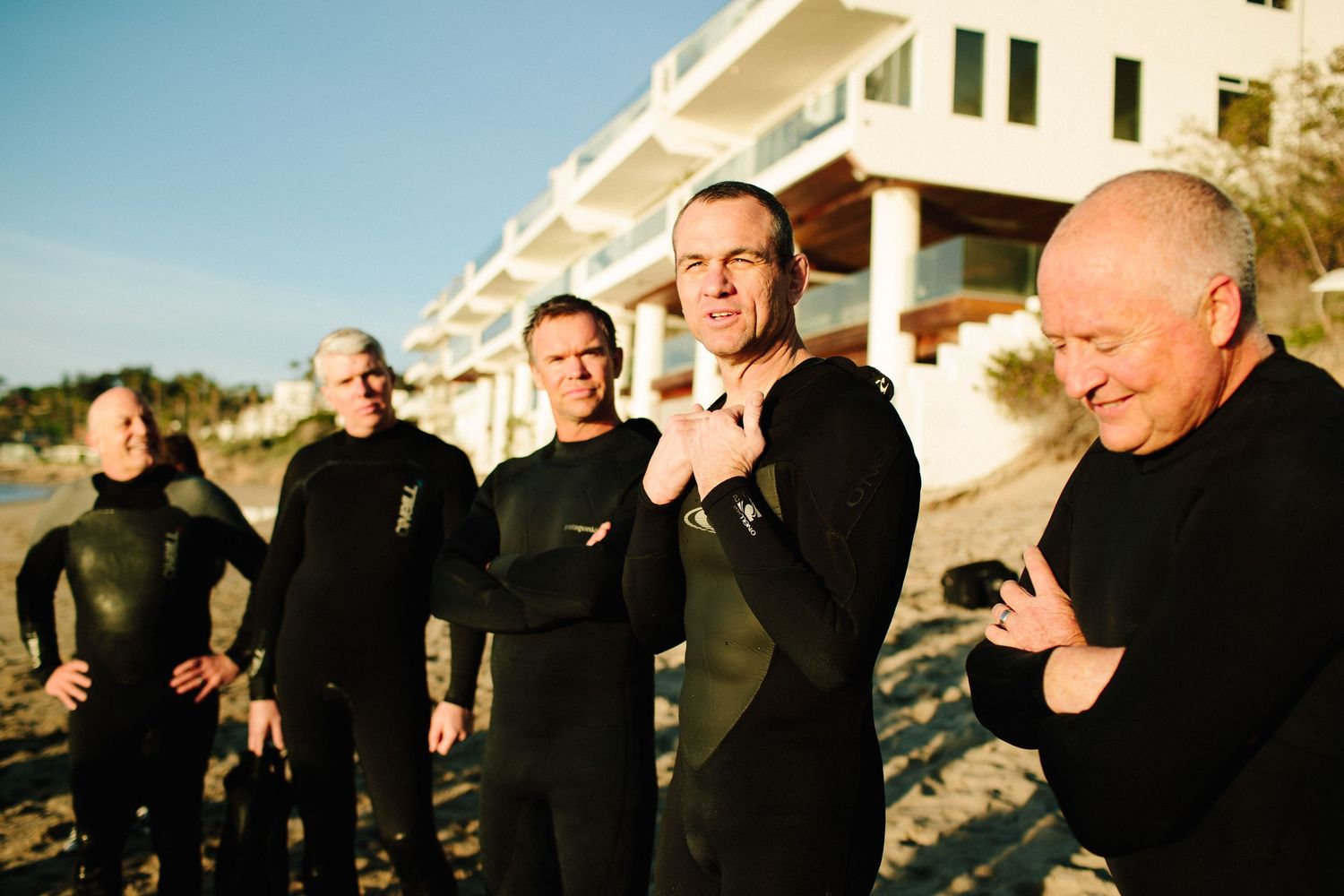 Group of people in dark clothing gathered outdoors near coastal homes during sunset.