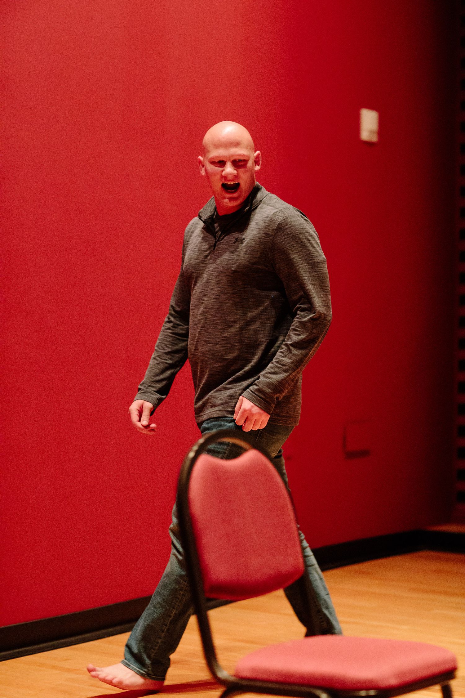 A performer in dark clothing moves energetically around a red chair on a wooden stage.