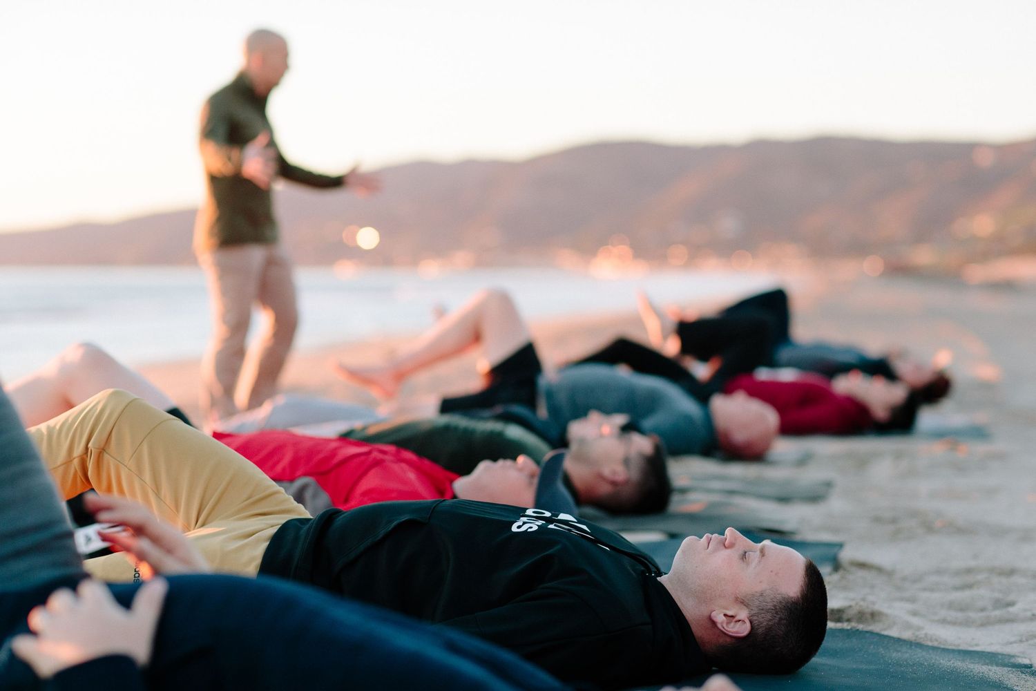 Participants lie on their backs doing relaxation exercises on a beach at sunset.
