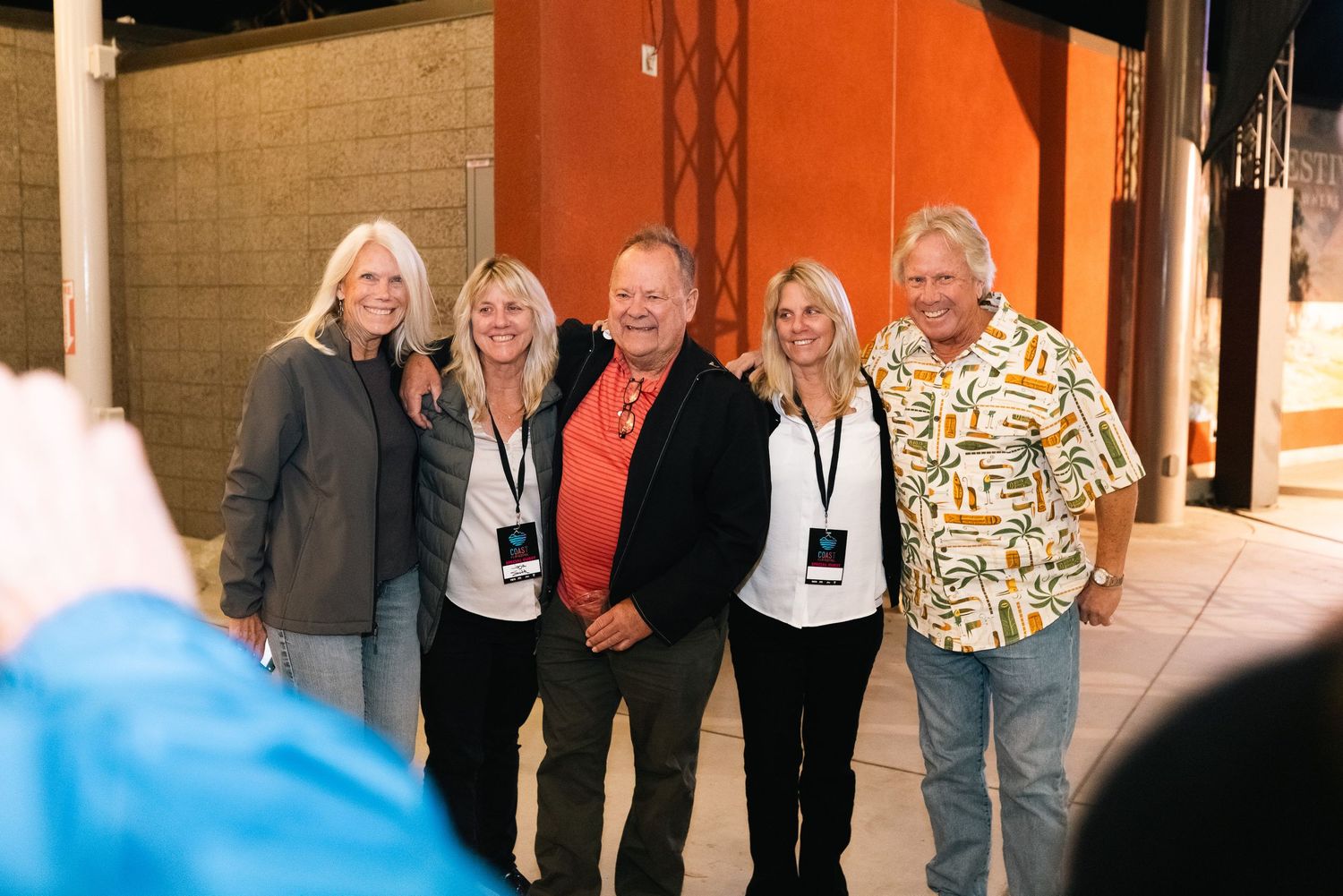 A group of people wearing event badges pose together outside a building with orange walls and columns.
