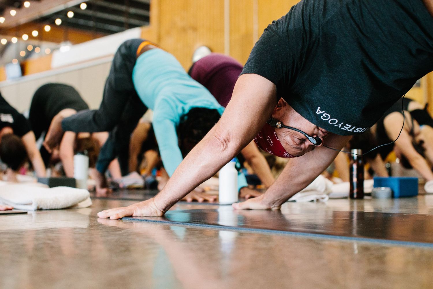 Close-up of yoga students doing downward dog pose on wooden floor with string lights above.