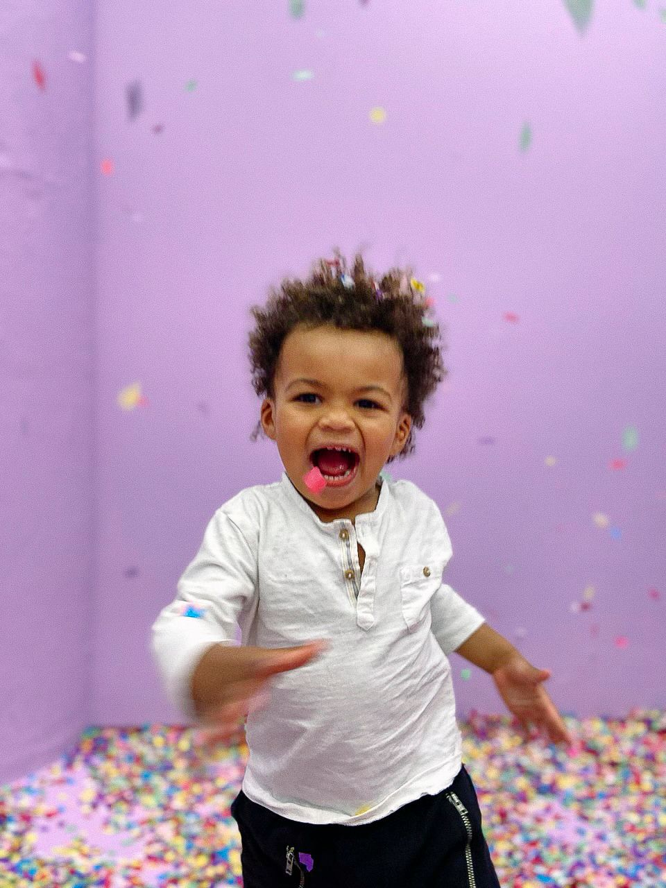 Excited small child in white shirt jumps and plays in a room with colorful confetti against a purple wall.