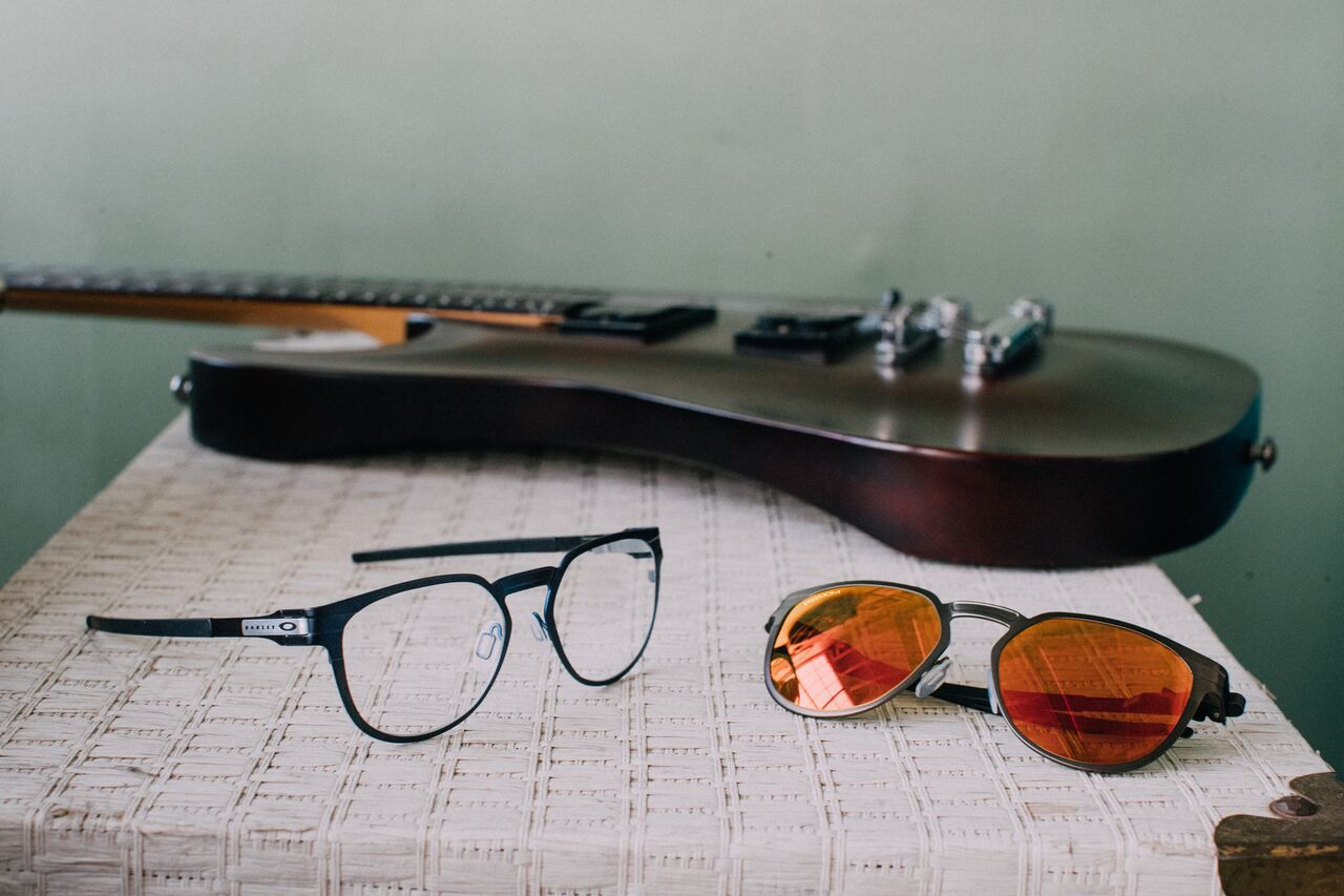 Black guitar, prescription glasses, and tinted sunglasses arranged on sheet music against a teal background.