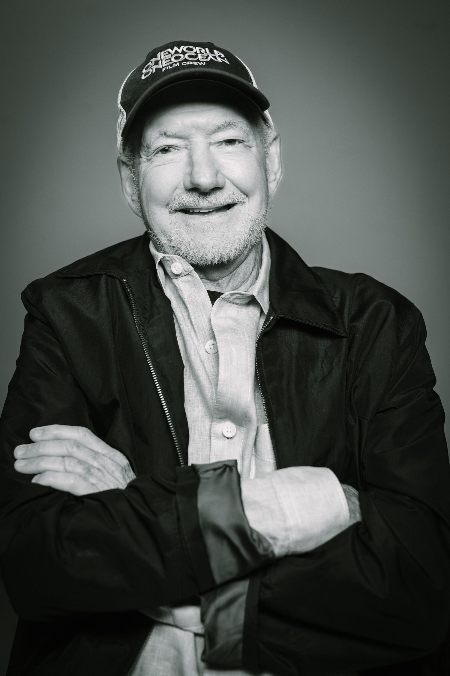 Black and white headshot of a senior man wearing a baseball cap and jacket with crossed arms.