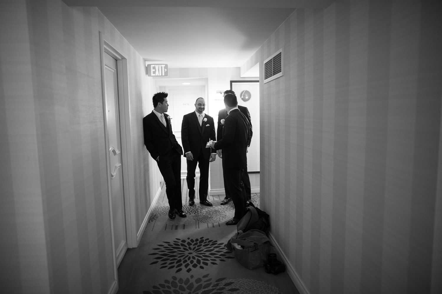 Three dark silhouettes stand in conversation at the end of a dimly lit hotel hallway.