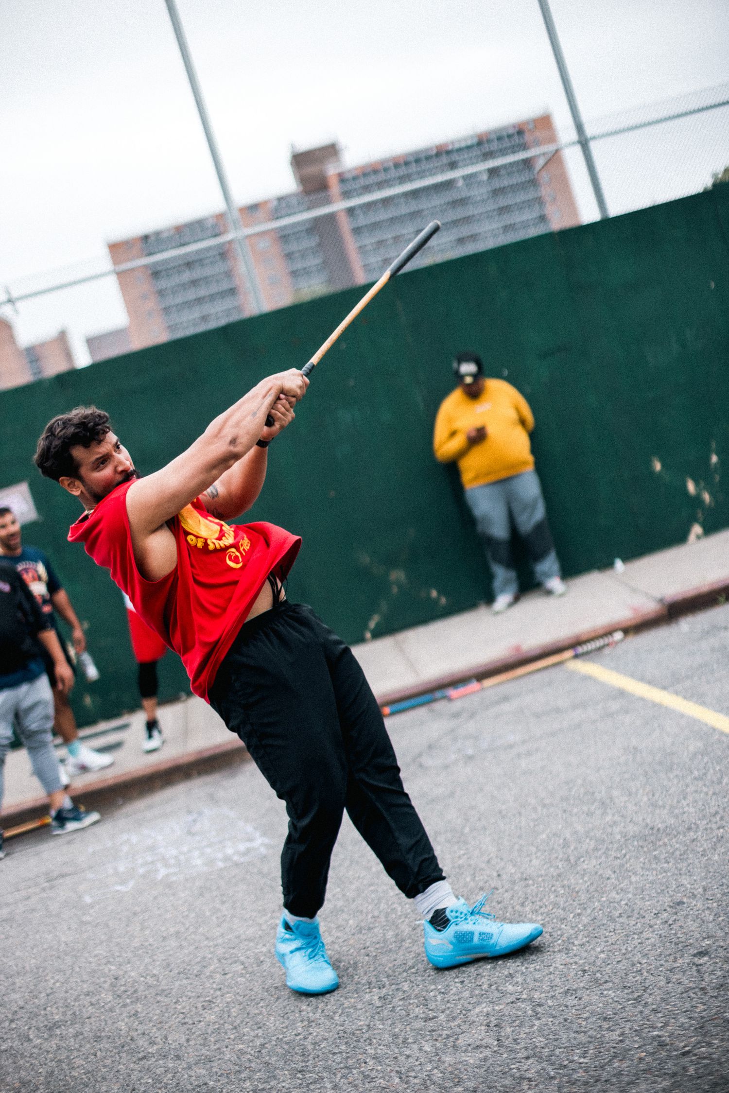 Player in red t-shirt demonstrates dramatic follow-through swing motion during urban stickball game.