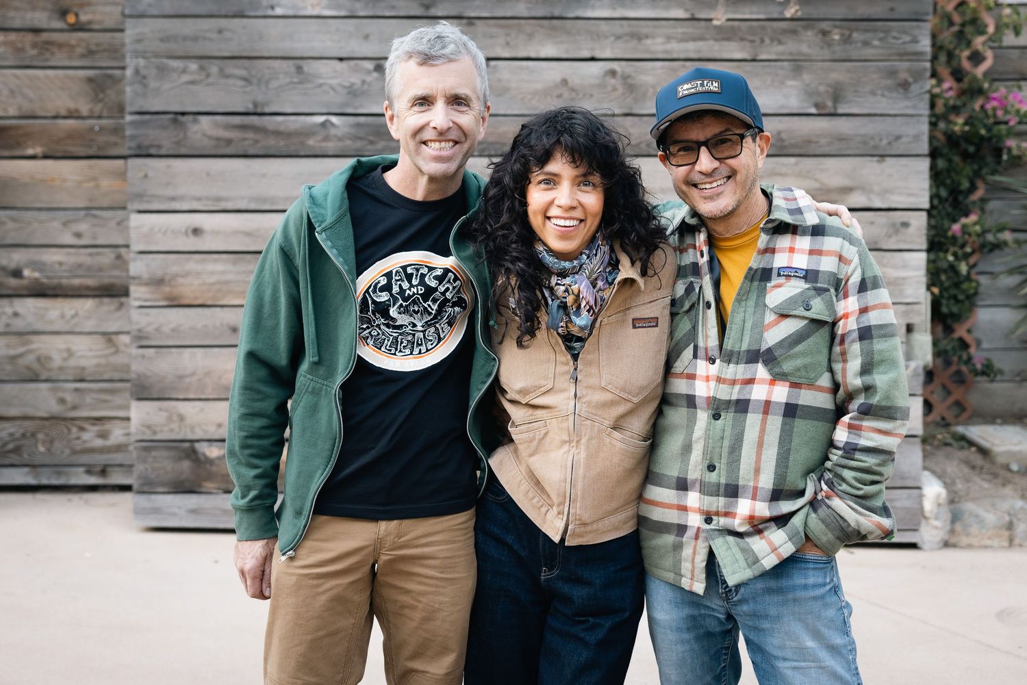 Three colleagues in casual wear smile together while standing in front of a rustic wooden wall.