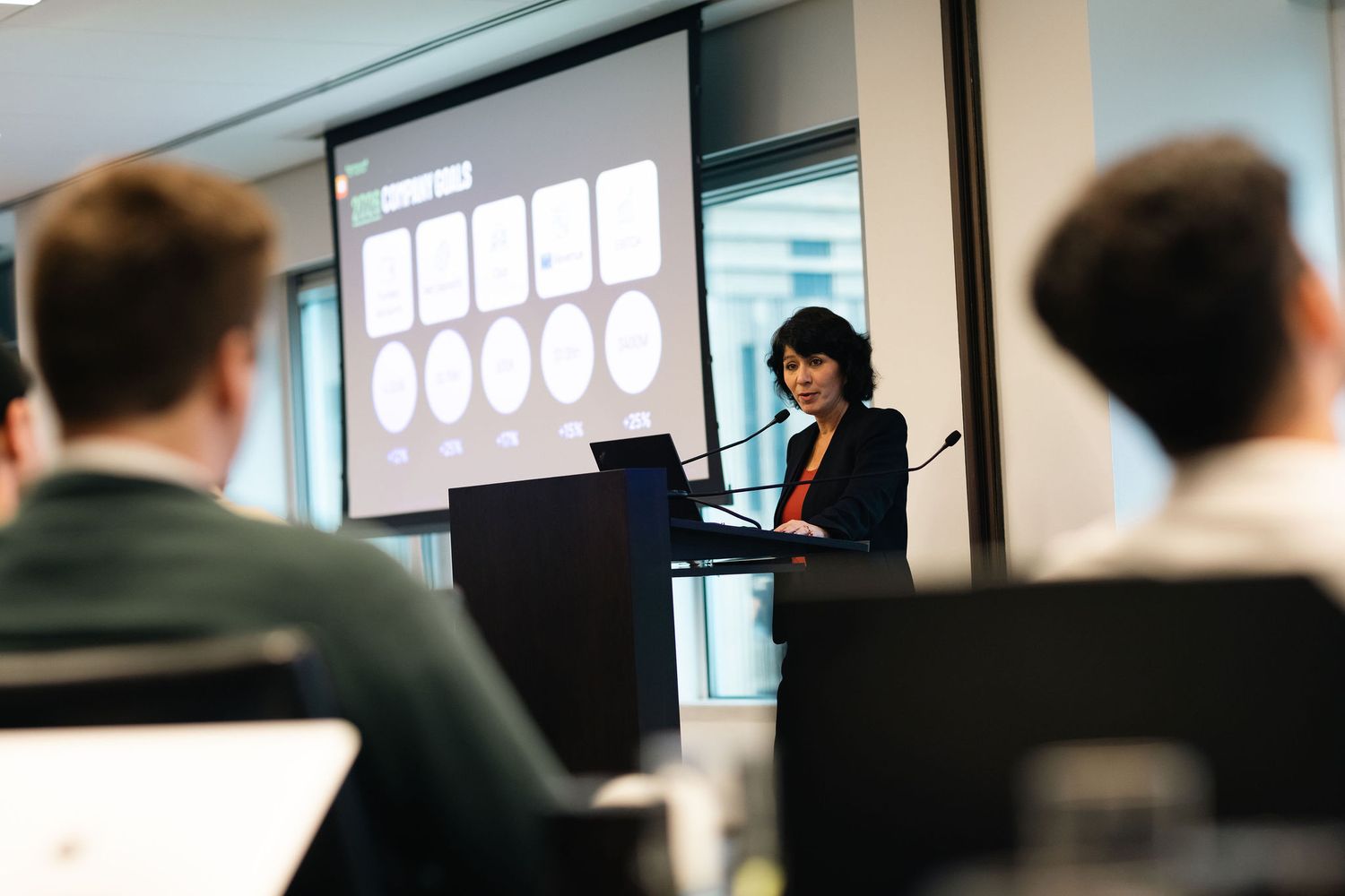 A presenter gives a business presentation in a conference room with a white projection screen visible in the background.