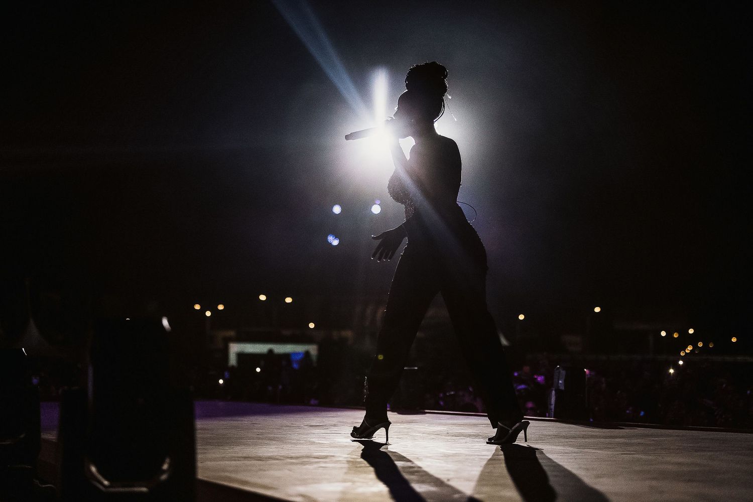 Silhouetted figures perform on a dark stage backlit by bright spotlights in a dramatic concert setting.