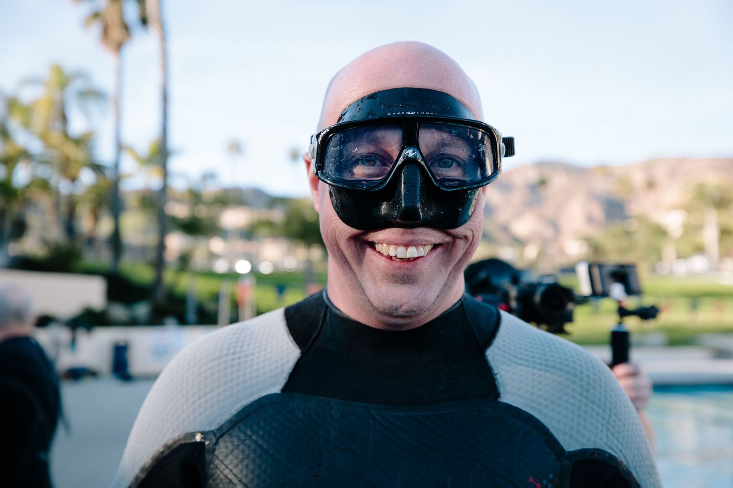 Person wearing protective goggles and diving gear smiling near a swimming pool.