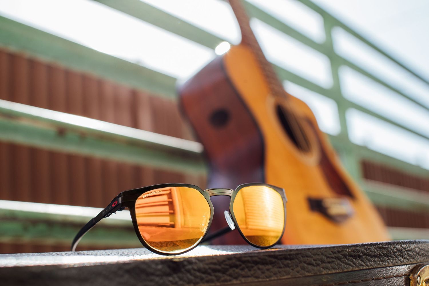 Sunglasses reflecting sunlight on wooden bench with acoustic guitar blurred in background.