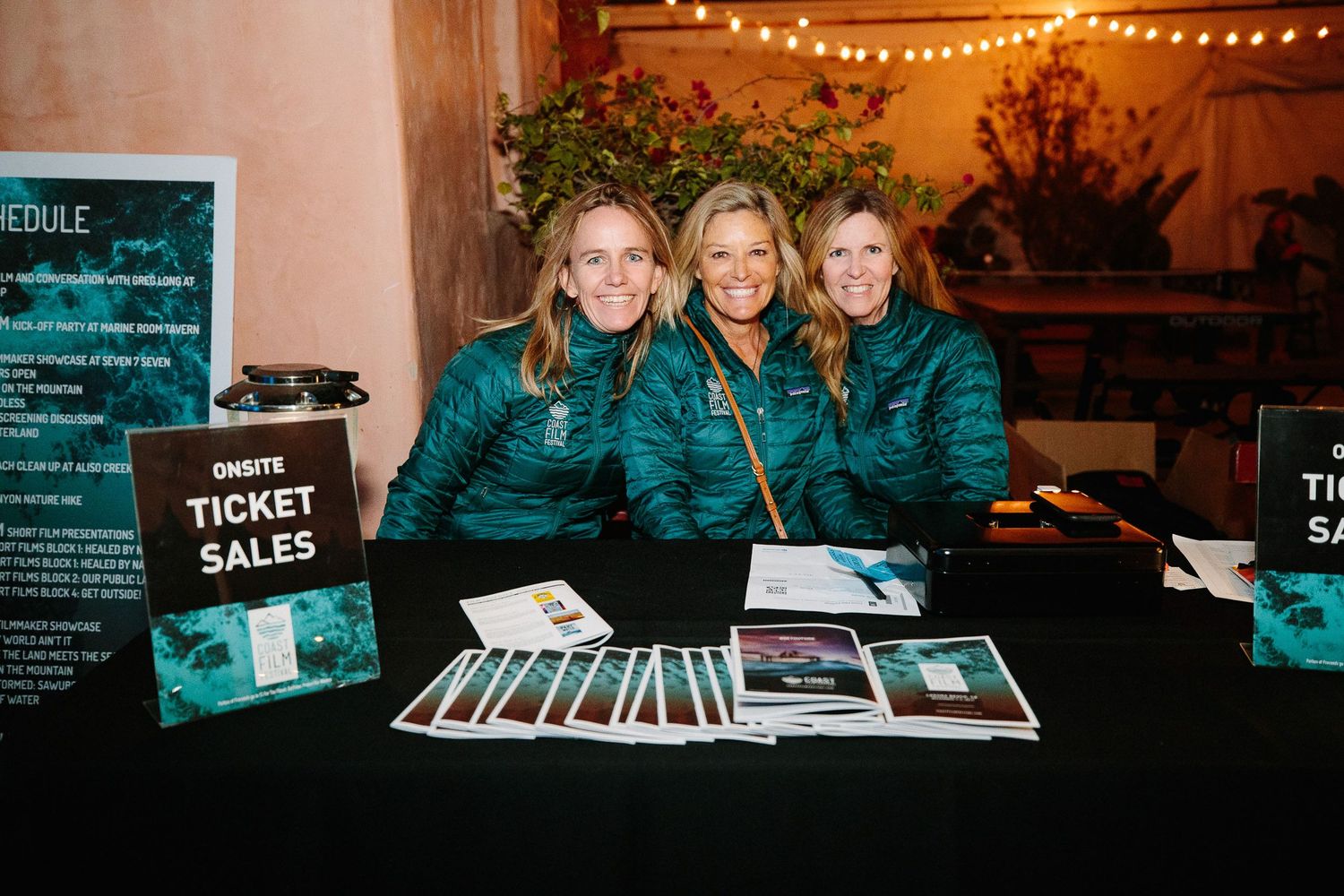 Three staff members in matching teal jackets manage a ticket sales booth at an event.