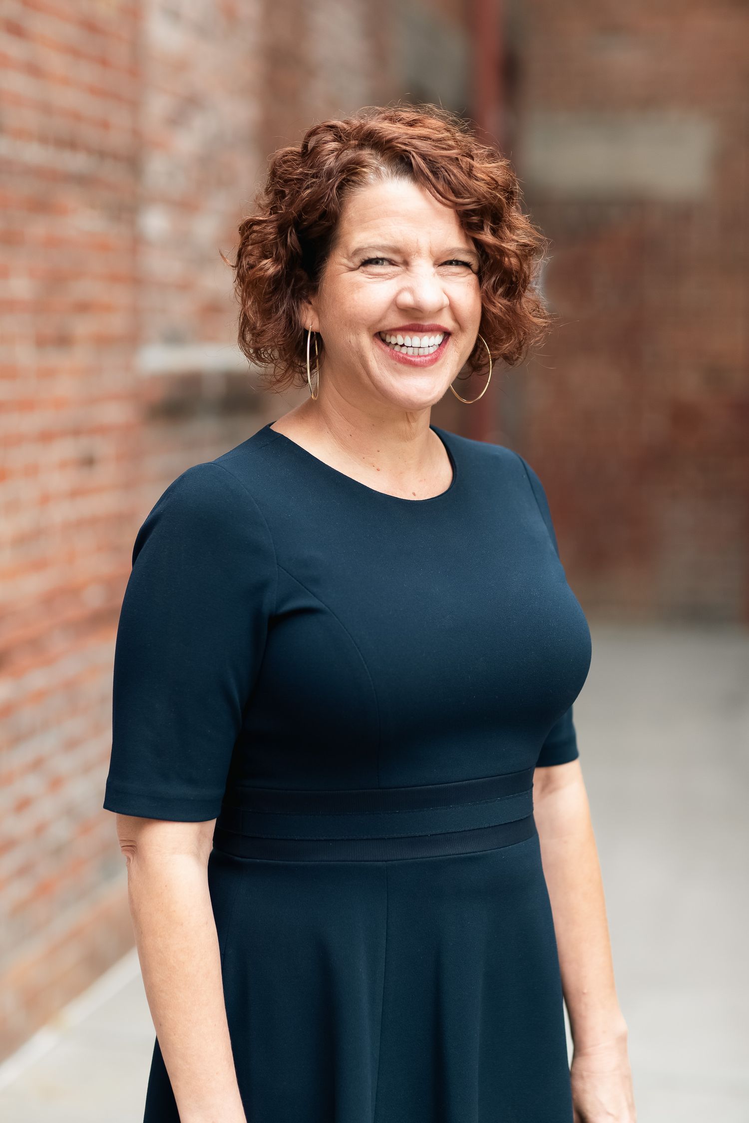 Woman in navy blue dress smiles while standing against brick wall background with short curly hair.