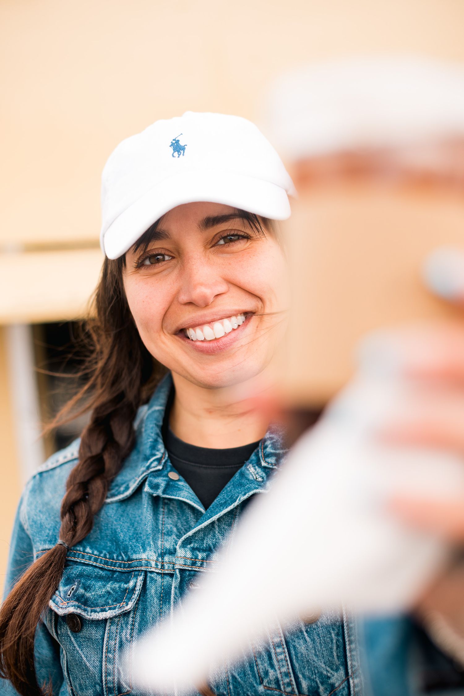 Woman in denim jacket and white baseball cap smiling brightly at camera.
