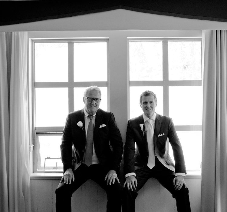 Formal group portrait of men in dark suits sitting against bright windows.