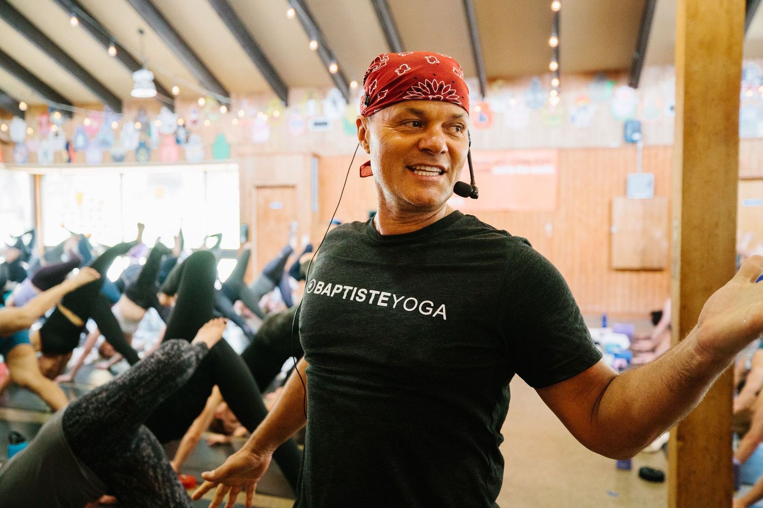 An energetic yoga instructor wearing a BaptisteYoga t-shirt and bandana leads a class.