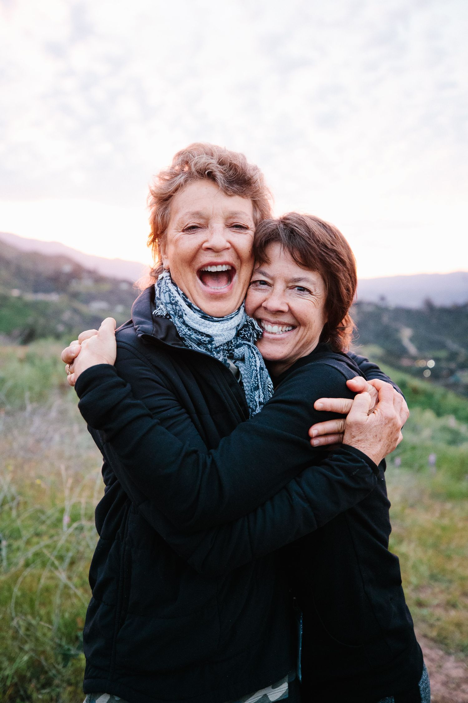 Two people embracing and laughing joyfully outdoors with mountains in the background.