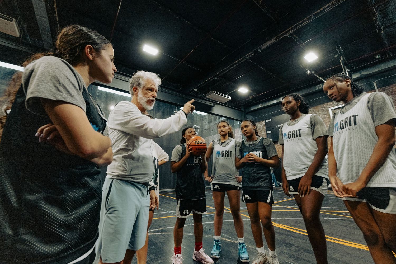 Basketball coach gives instruction to team in huddle during practice session on indoor court.