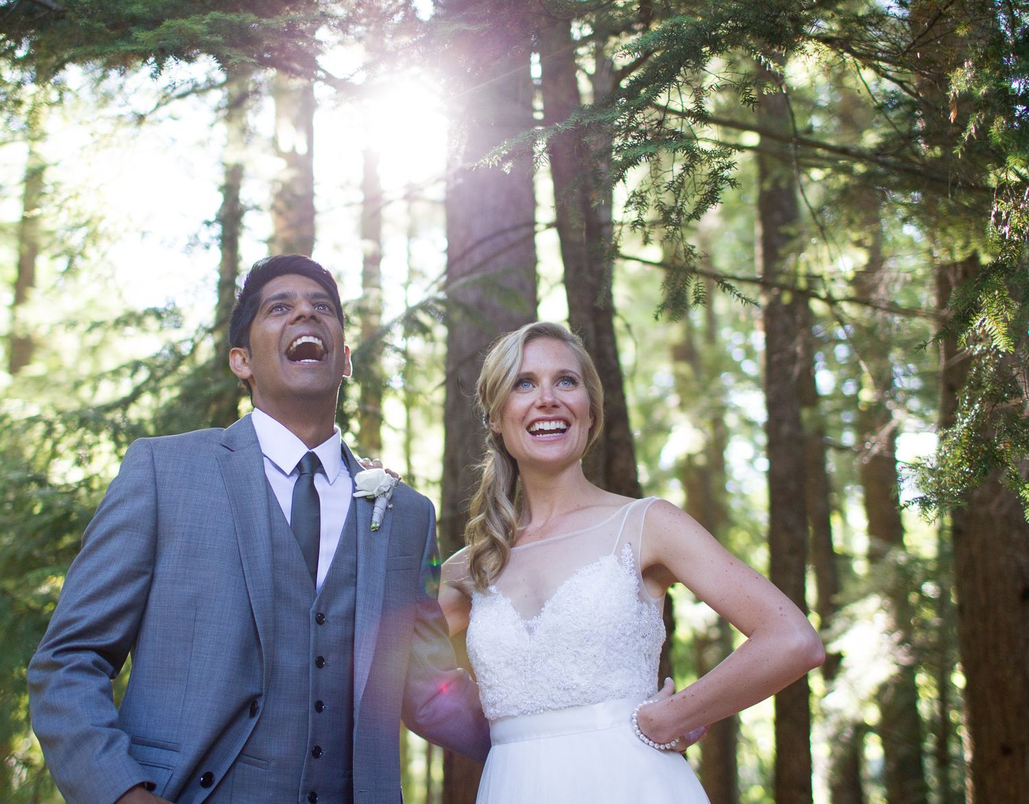 A joyful wedding couple laughing together in a sunlit forest setting.