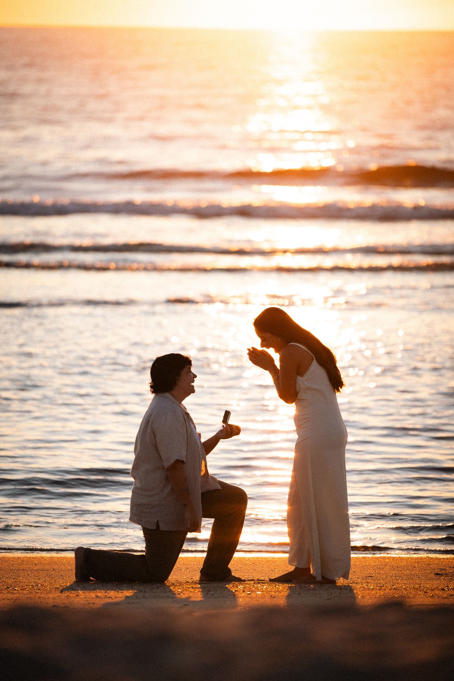 Two silhouettes sit together on a beach at sunset while waves gently roll onto the shore in golden light.