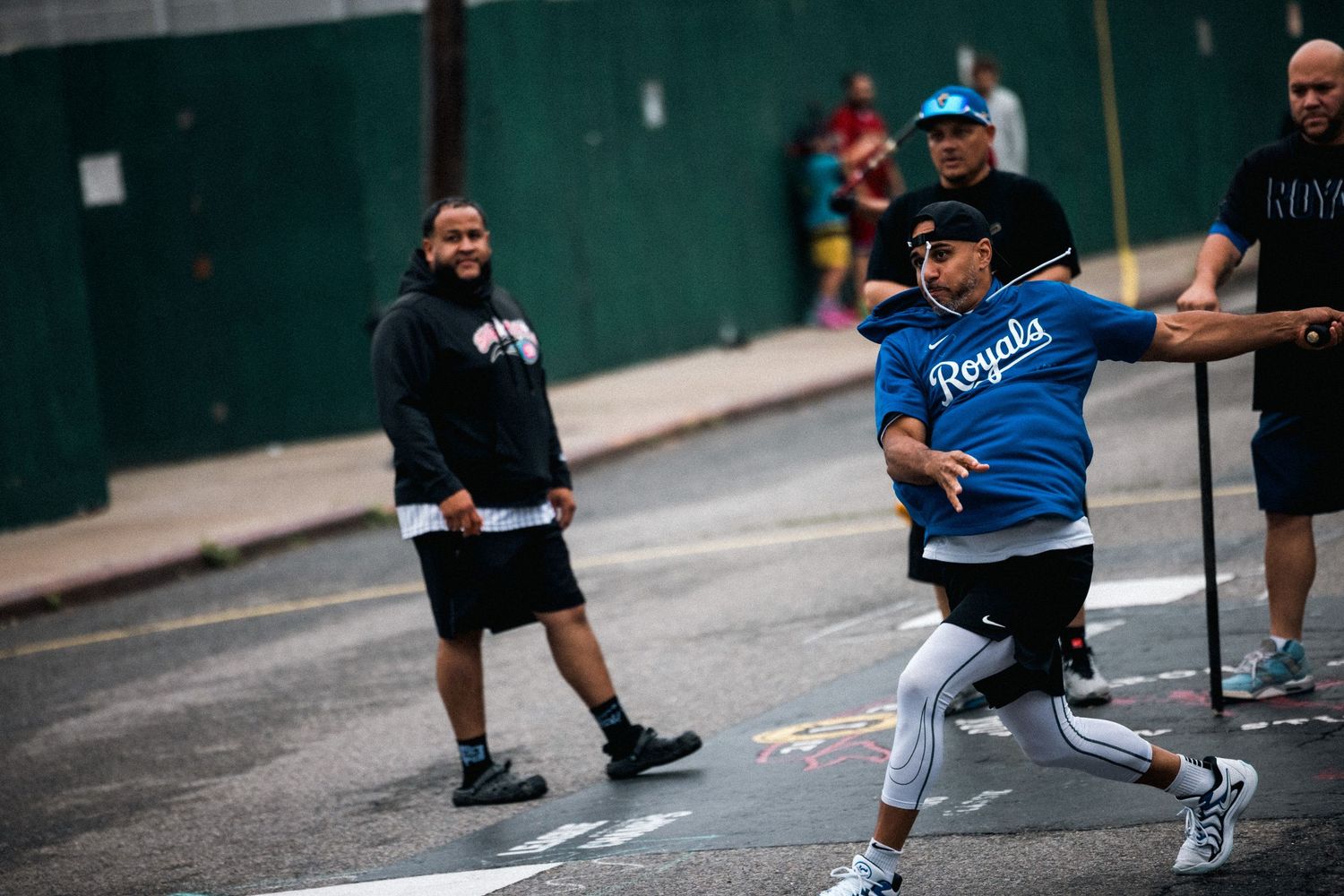 Dancers perform breakdancing moves on pavement in front of a green wall.