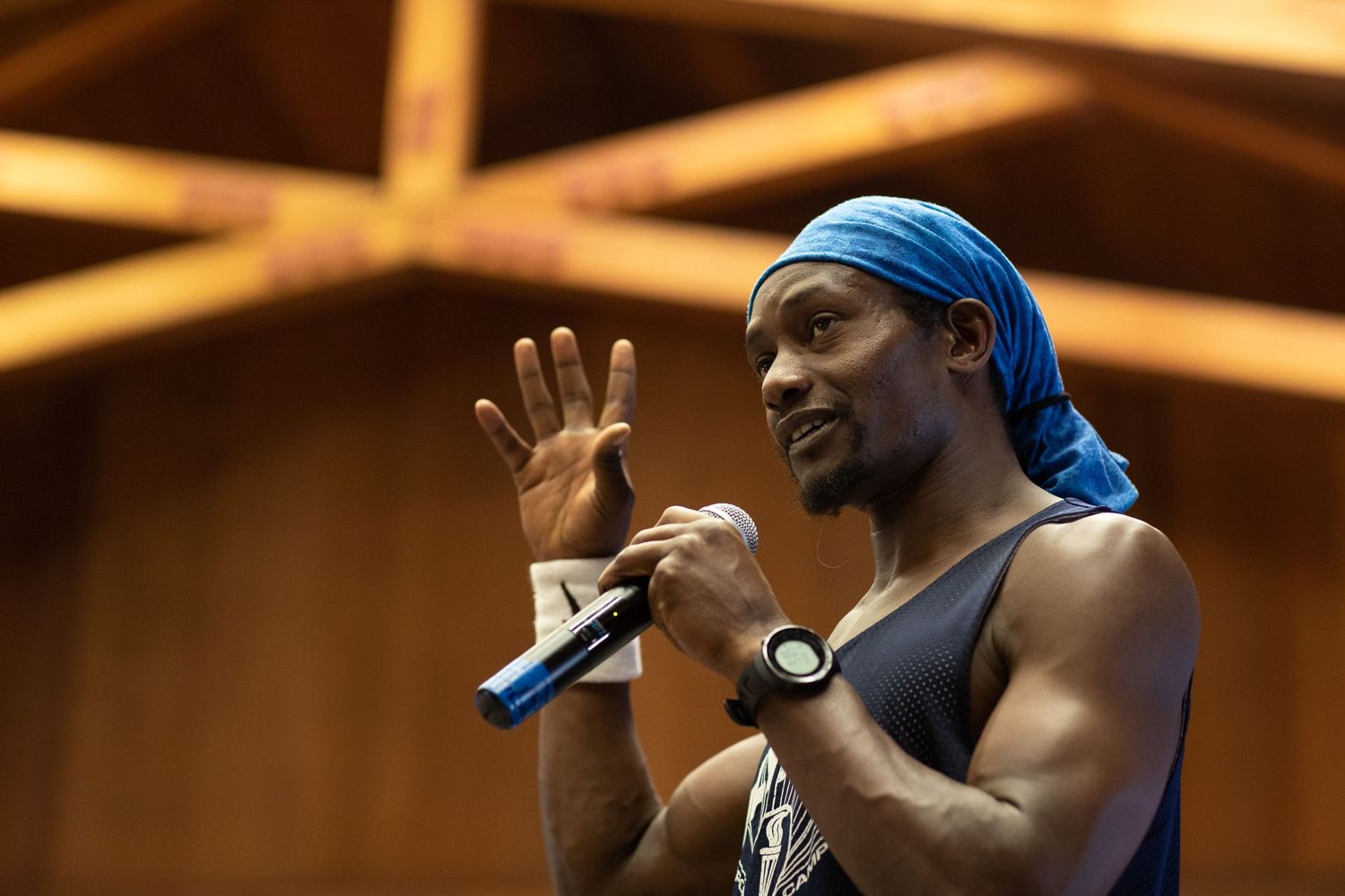 Fitness instructor in blue headband speaks into microphone during indoor presentation.